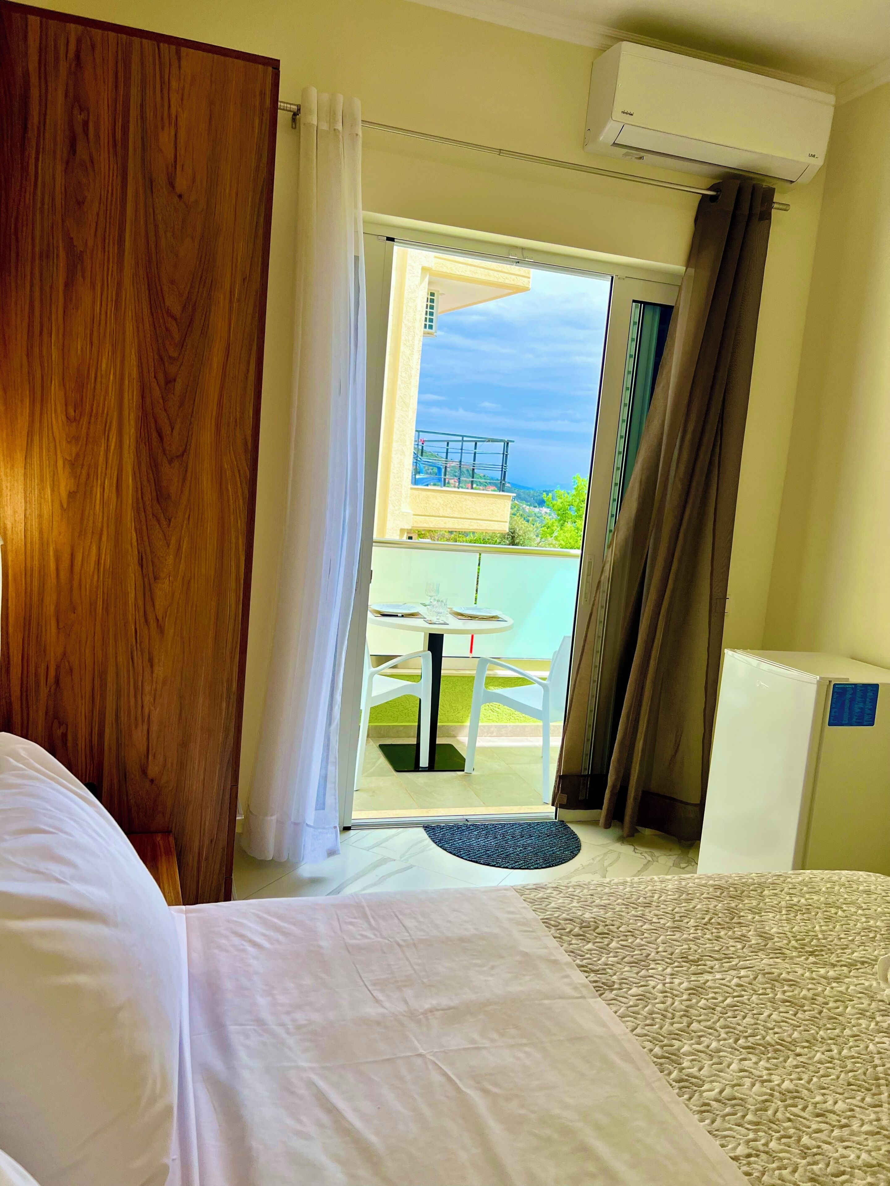 A hotel room with a bed, a desk, a chair, a small table with a chair, and a view of a balcony with a table and chairs.