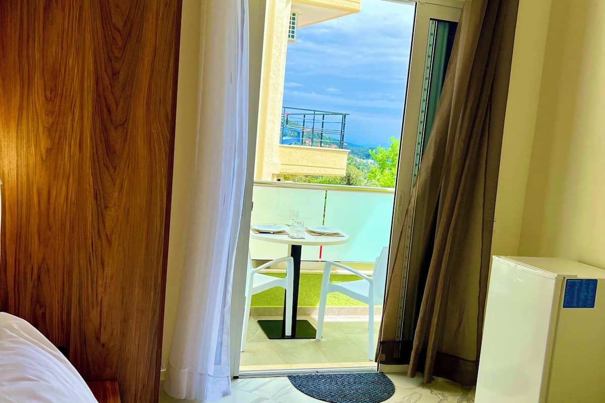 Una habitación de hotel con una cama, un escritorio, una silla, una mesita con otra silla, y vistas a un balcón con mesa y sillas.