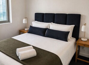 Cama bem arrumada com cabeceira escura, roupa de cama branca e duas luminárias sobre mesas de cabeceira de madeira.