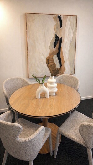 Una mesa de madera redonda con dos jarrones blancos y una planta, rodeada por cuatro sillas con estampado, con un cuadro abstracto grande en la pared.
