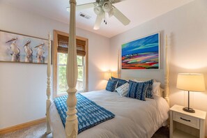 A bedroom with a four-poster bed, a ceiling fan, a nightstand with a lamp, and artwork on the walls.