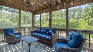 A covered outdoor seating area with wicker furniture, ceiling fans, and large windows offering a forest view.