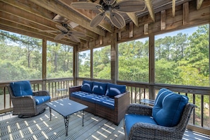A covered outdoor seating area with wicker furniture, ceiling fans, and large windows offering a forest view.