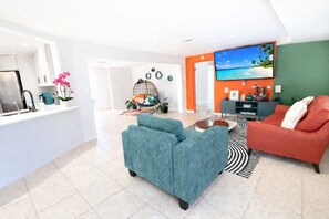 A modern living room with a sofa, a coffee table, and a TV. There is a kitchen area with a fridge and a sink.