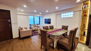 A modern living room with a glass-top dining table, brown upholstered chairs, a sofa, and a TV mounted on the wall. - A fully furnished, modern, comfortable, and cozy apartment. (Cusco)