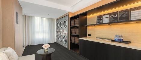 Una habitación de hotel con sofá, una pequeña mesa redonda con un florero, zona de lavandería con lavadoras y secadoras, y una recepción con paneles informativos.