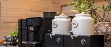 A coffee station with a coffee machine, two large containers, and a decorative plant.