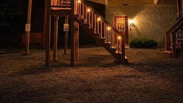 Una casa de madera de dos pisos con una escalera que lleva a un balcón, una bandera y un exterior bien iluminado.