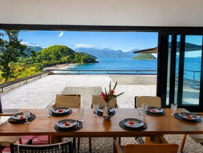A dining table with plates and glasses, set against a scenic view of a beach and mountains.