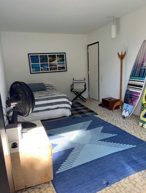 Une chambre avec un lit, un ventilateur sur pied, une chaise, une planche de surf et une photo encadrée accrochée au mur.