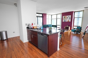 A modern kitchen with a dishwasher, a sink, and a countertop. There is a dining area with a teal sofa and a small table. The room has large windows and a view of the city.