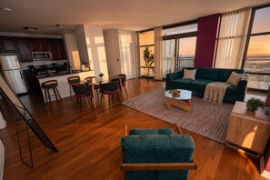Un soggiorno moderno con divano, tavolino e poltrone. È presente una zona cucina con frigorifero, microonde e fornello. Ampie finestre offrono vista sulla città.