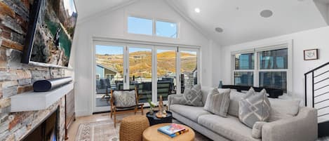 A modern living room with a stone fireplace, a grey sofa, a round wooden coffee table, and a large TV mounted on the wall.