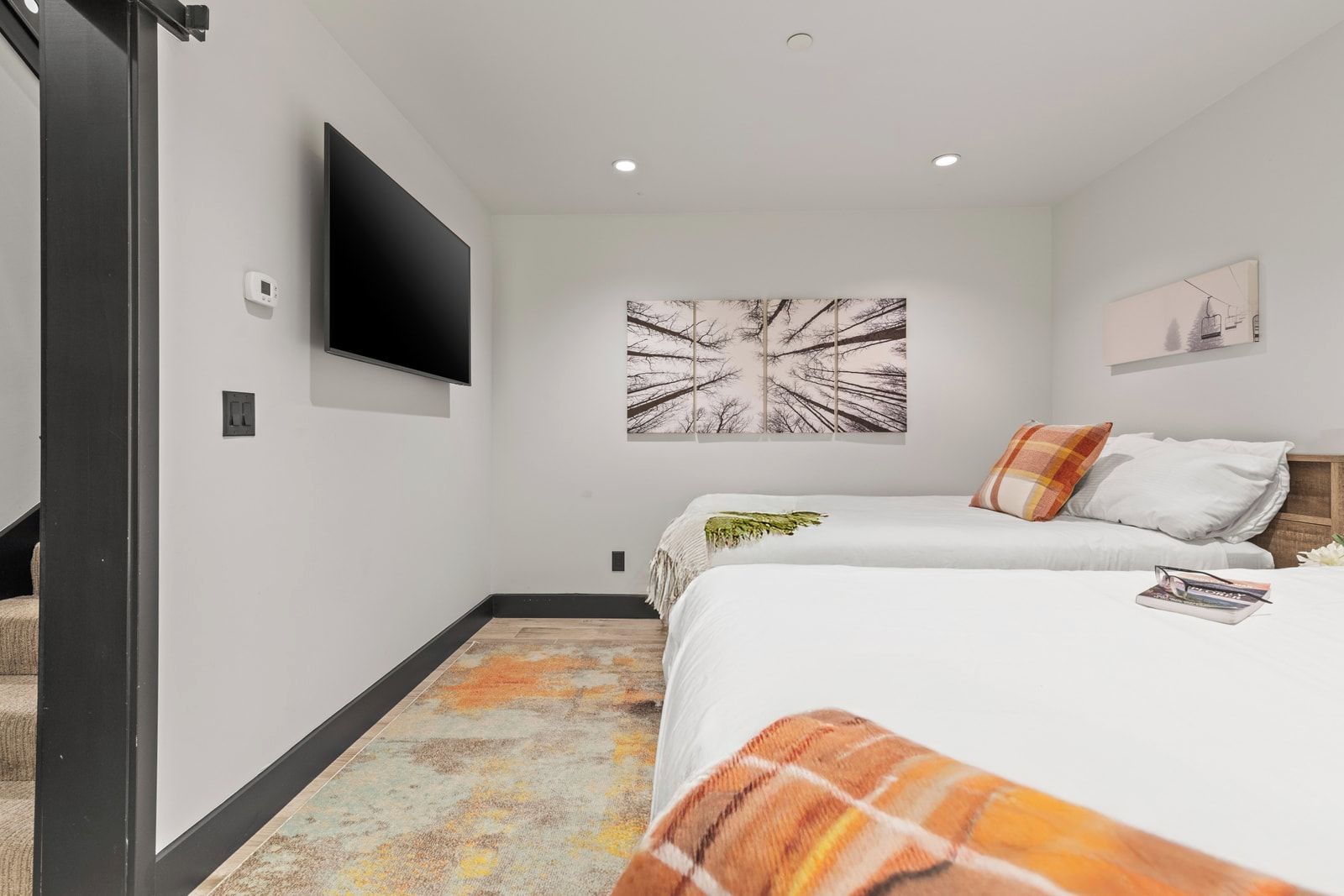A hotel room with two beds, a flat-screen TV, a painting, and a small shelf with a book.