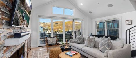 A modern living room with a stone fireplace, a grey sofa, a round wooden coffee table, and a large TV mounted on the wall.