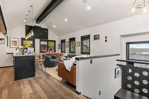 A modern living room with a dining area, kitchen, and a large window with a view. - Walk to Ski Slopes + Trails! 2 Mountain View Homes, 7 Bedrooms, with Private Hot Tubs, Fireplaces, and AC | ML-3981 by KBM (Park City)