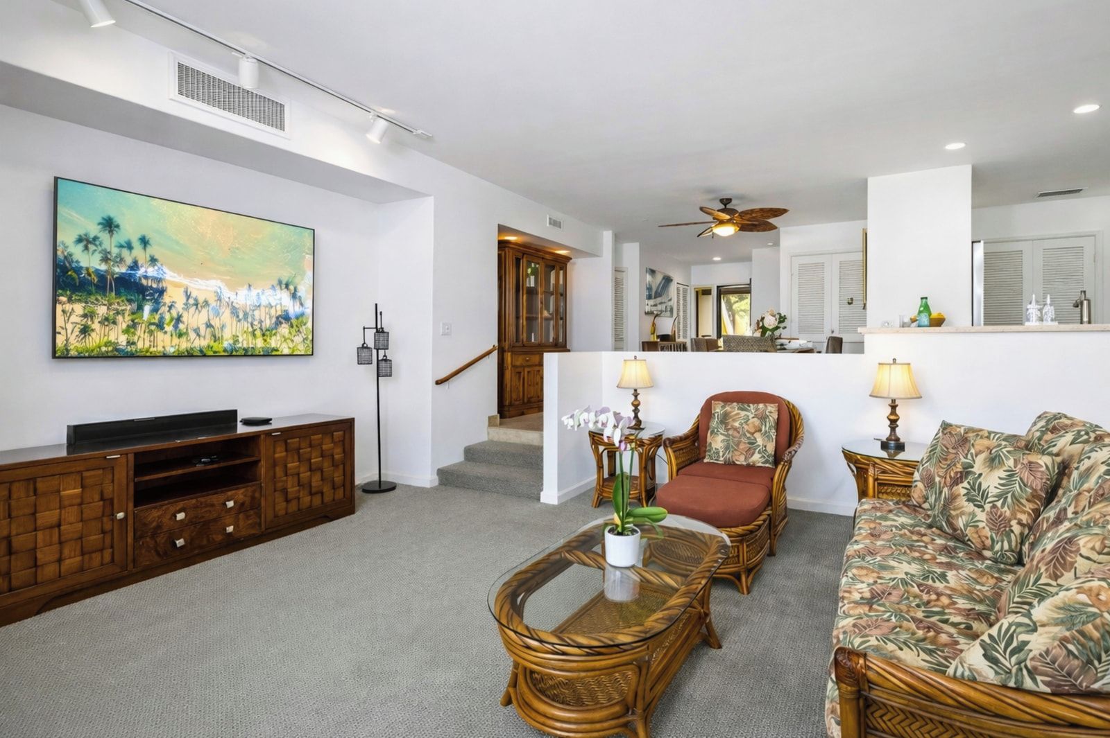 A spacious living room with a flat-screen TV, a large painting, wicker furniture, and a staircase leading to a kitchen area.