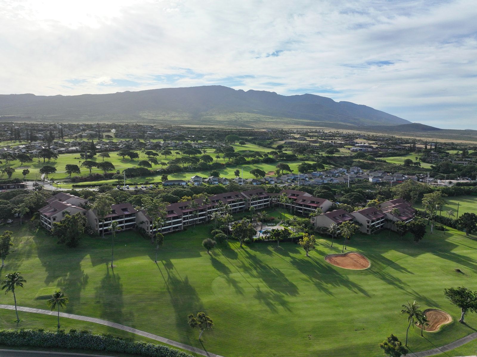 A golf course with a hotel complex in the foreground, surrounded by greenery and mountains in the background.