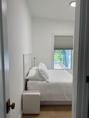 A bedroom with a bed, a nightstand, a window with blinds, and a door. - Modern Beach Cottage (Sag Harbor)