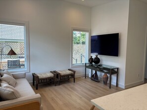 A modern living room with a sofa, two chairs, a glass coffee table, a TV mounted on the wall, and large windows with blinds. - Modern Beach Cottage (Sag Harbor)