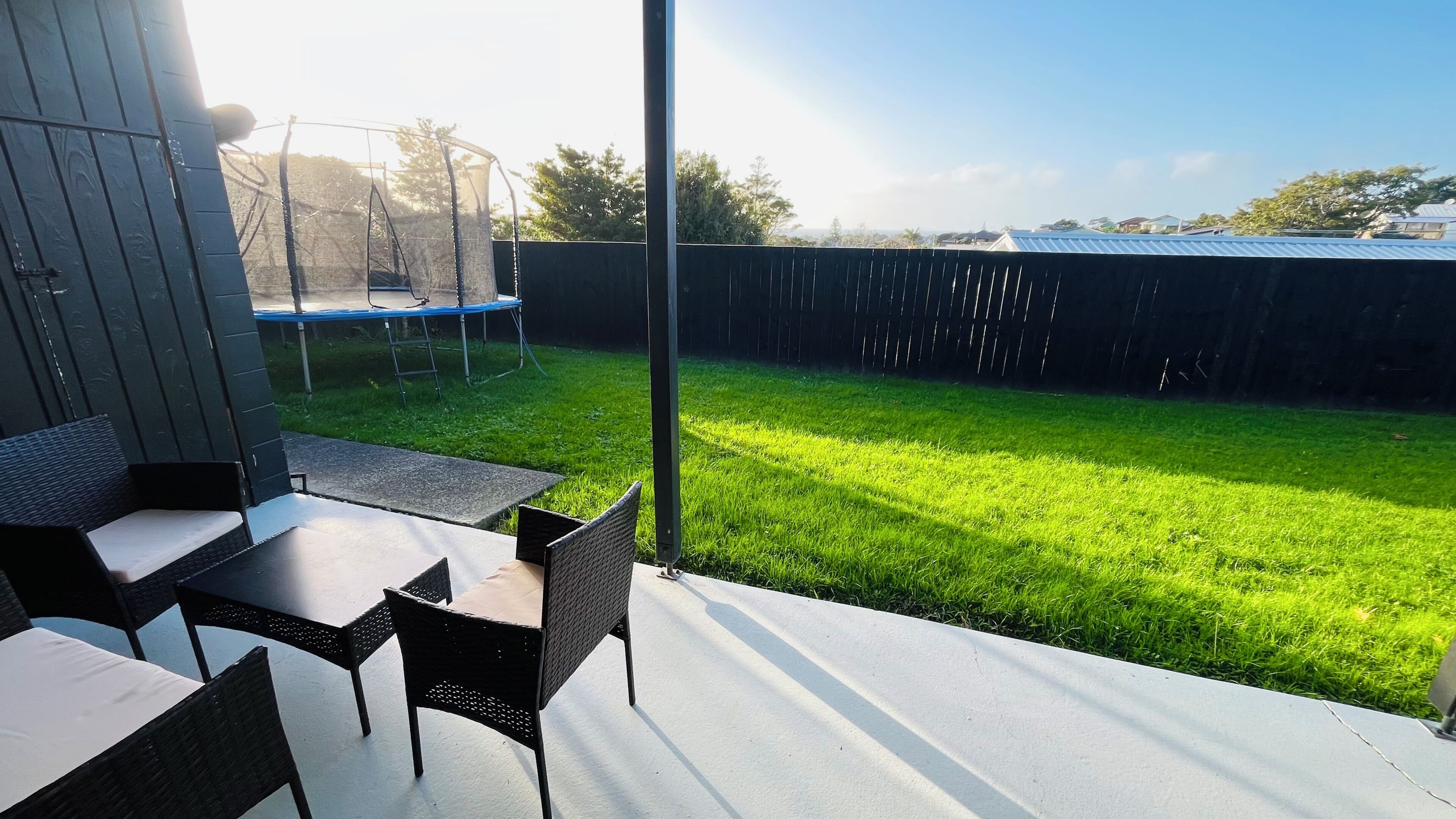 Un patio con mobiliario exterior, un trampolín y vista a un jardín cubierto de césped.