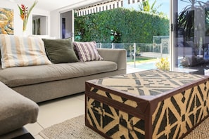 A modern living room with a sofa, a patterned ottoman, and a view of a well-maintained garden through a glass door.