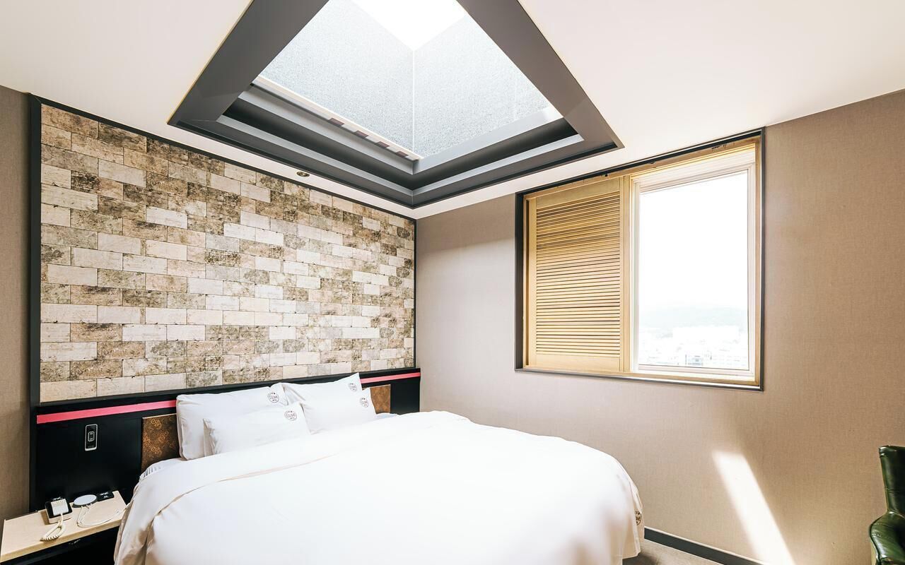 A hotel room with a skylight, a brick-patterned headboard, a bed, a bedside table with a phone, and a window with blinds.