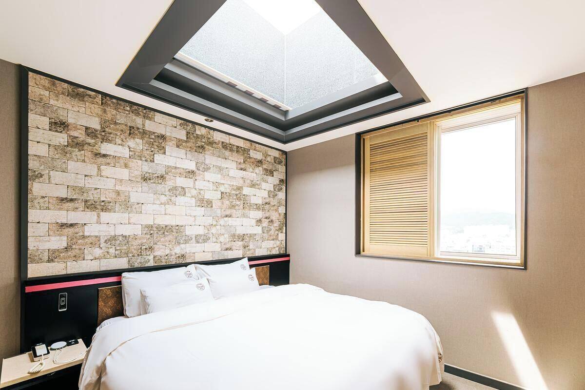 Una habitación de hotel con tragaluz, cabecera estampada con diseño de ladrillo, cama, mesita de noche con teléfono y ventana con persianas.