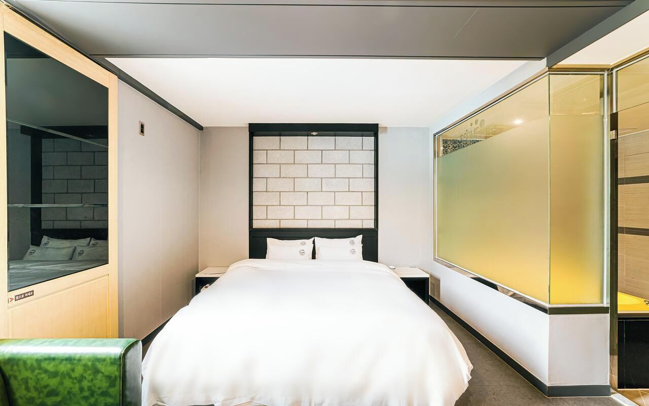A modern hotel room with a large bed, a nightstand, a glass partition, and a green chair.