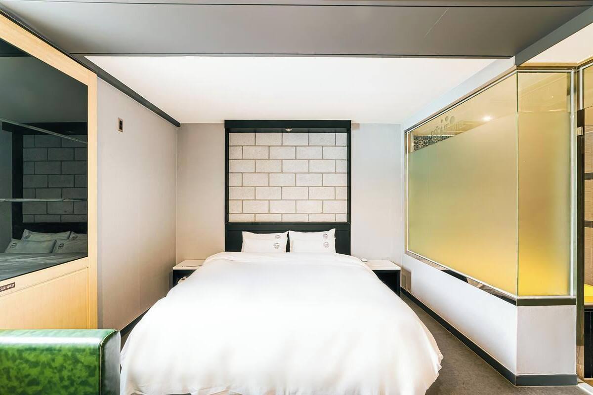 Una habitación de hotel moderna con una cama grande, una mesita de noche, un separador de vidrio y una silla verde.