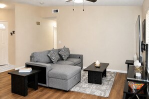 A living room with a gray sectional sofa, two black and white patterned pillows, a black coffee table, a black side table, and a TV on a black stand.