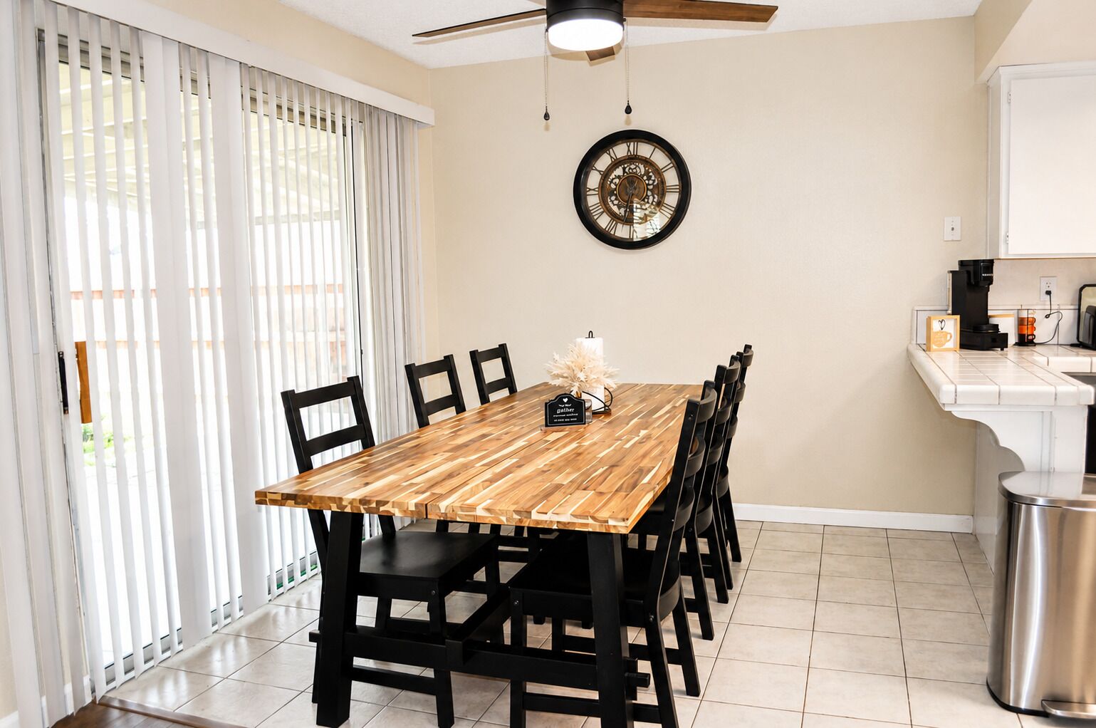 A dining area with a wooden table, black chairs, a wall clock, and a kitchen counter with a coffee machine.