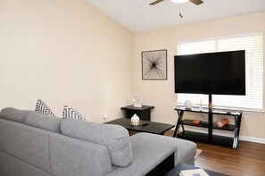 Una sala de estar con un sofá gris, un televisor de pantalla plana, una mesa de centro y un objeto decorativo sobre un pedestal.