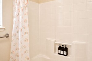Un baño con azulejos blancos, una cortina de ducha con motivos de arcoíris y tres botellas de artículos de aseo en un estante.