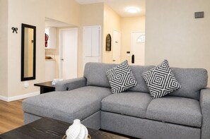A grey sectional sofa with patterned pillows, a small table, and a vase on a wooden coffee table in a well-lit room with beige walls and a door in the background. - Roomy 3BR & 2BA Retreat with Laundry & King Bed (Hanford)