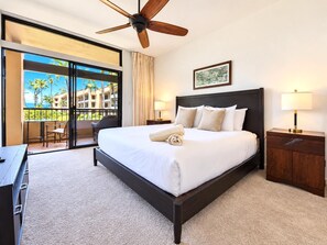 Quarto de hotel com cama grande, ventilador de teto, vista para palmeiras e um edifício através da porta de vidro, e escrivaninha com cadeira.