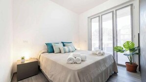 Un dormitorio con cama, almohadas, una mesita de noche, una planta en maceta y una ventana.