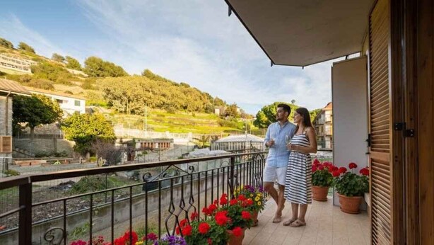 Un balcón con flores en macetas, vistas a un paisaje montañoso y un edificio al fondo.