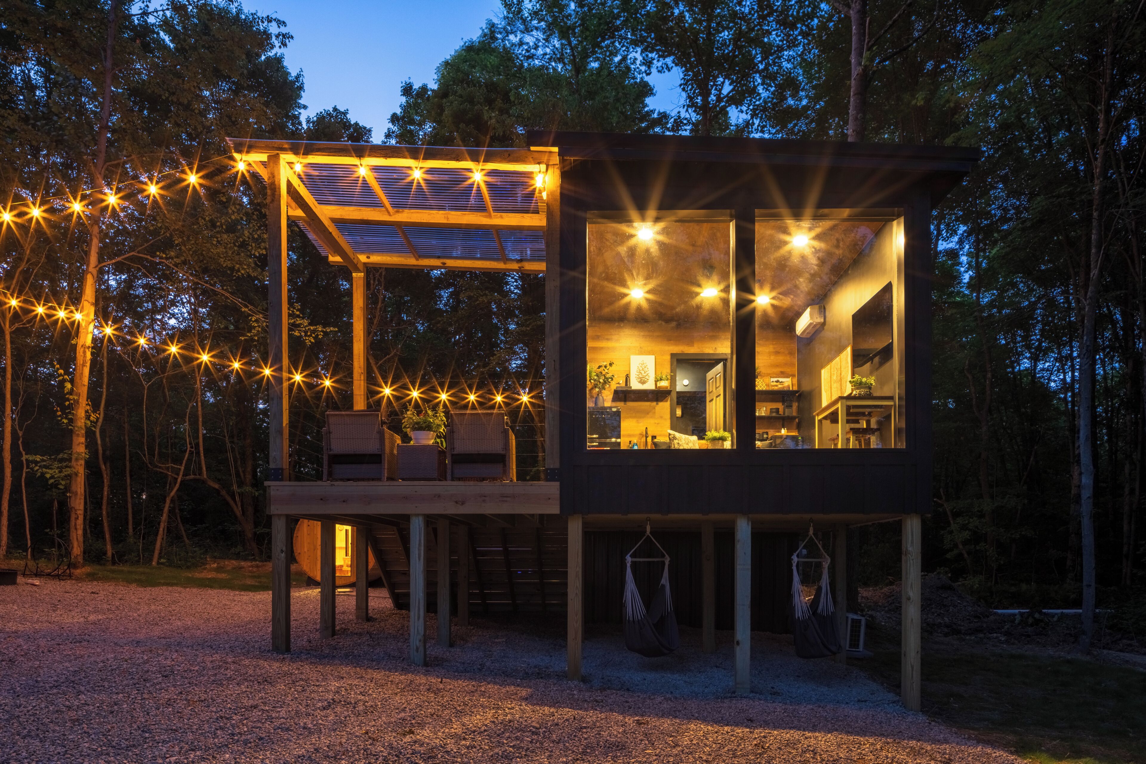 A modern wooden cabin with a glass-enclosed dining area, a deck with seating, and a hanging hammock.