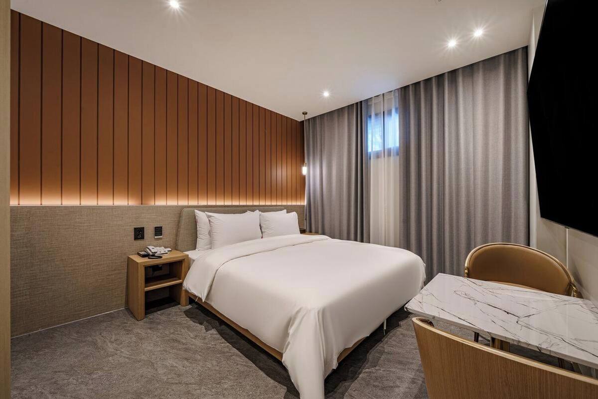 A modern hotel room with a large bed, a desk with a chair, a flat-screen TV, and a wooden panel wall.