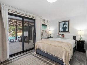 Un dormitorio con cama, mesita de noche, lámpara, un cuadro y vistas a una piscina exterior.