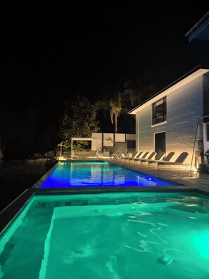 Una piscina iluminada con luz azul, una casa moderna con fachada de baldosas y algunas sillas de relax.