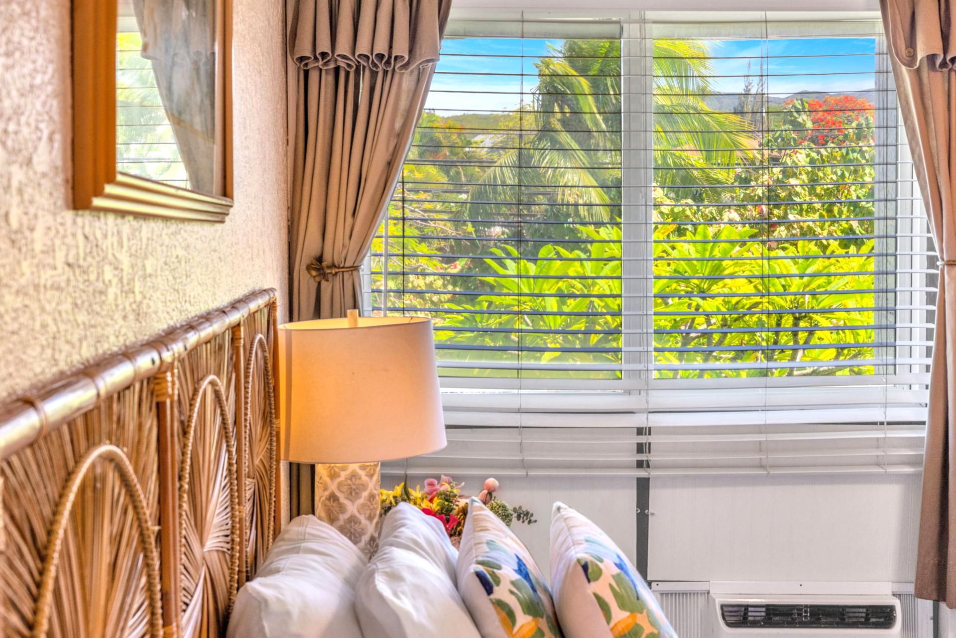 A hotel room with a bed, a bedside lamp, a window with blinds, and a view of greenery outside.