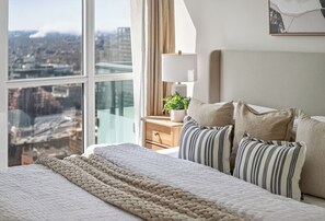 Ein Schlafzimmer mit großem Fenster, Nachttischlampe, Pflanze und einem Bett mit gestreifter Bettwäsche.