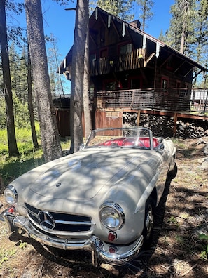 A vintage car parked in a forested area with a cabin in the background.