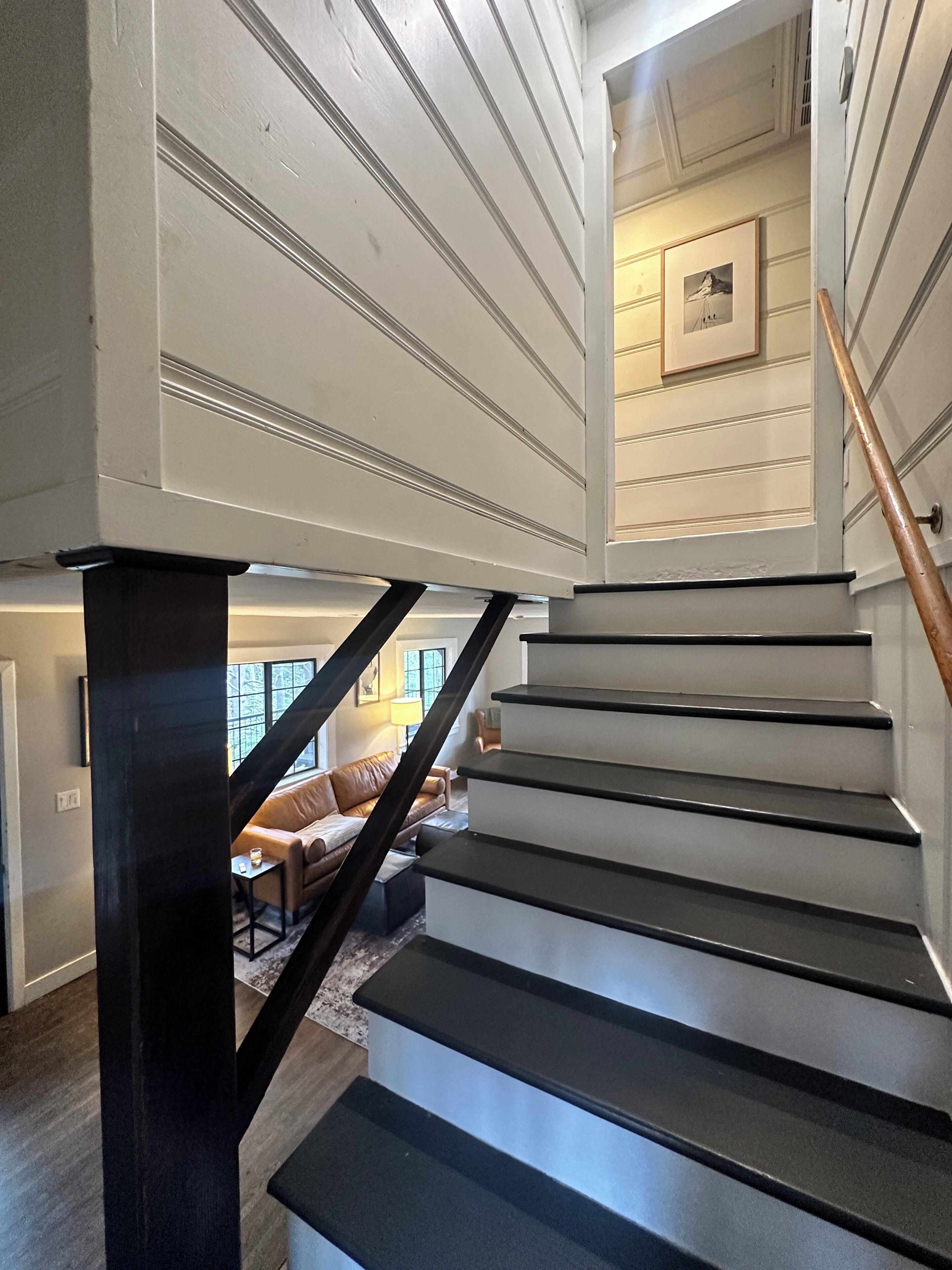 Uma escadaria com degraus pretos e corrimão de madeira, que dá acesso a uma sala de estar com sofá de couro e luminária.