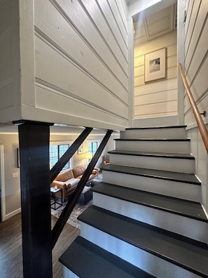 A staircase with black steps and a wooden handrail, leading to a living area with a leather couch and a lamp.