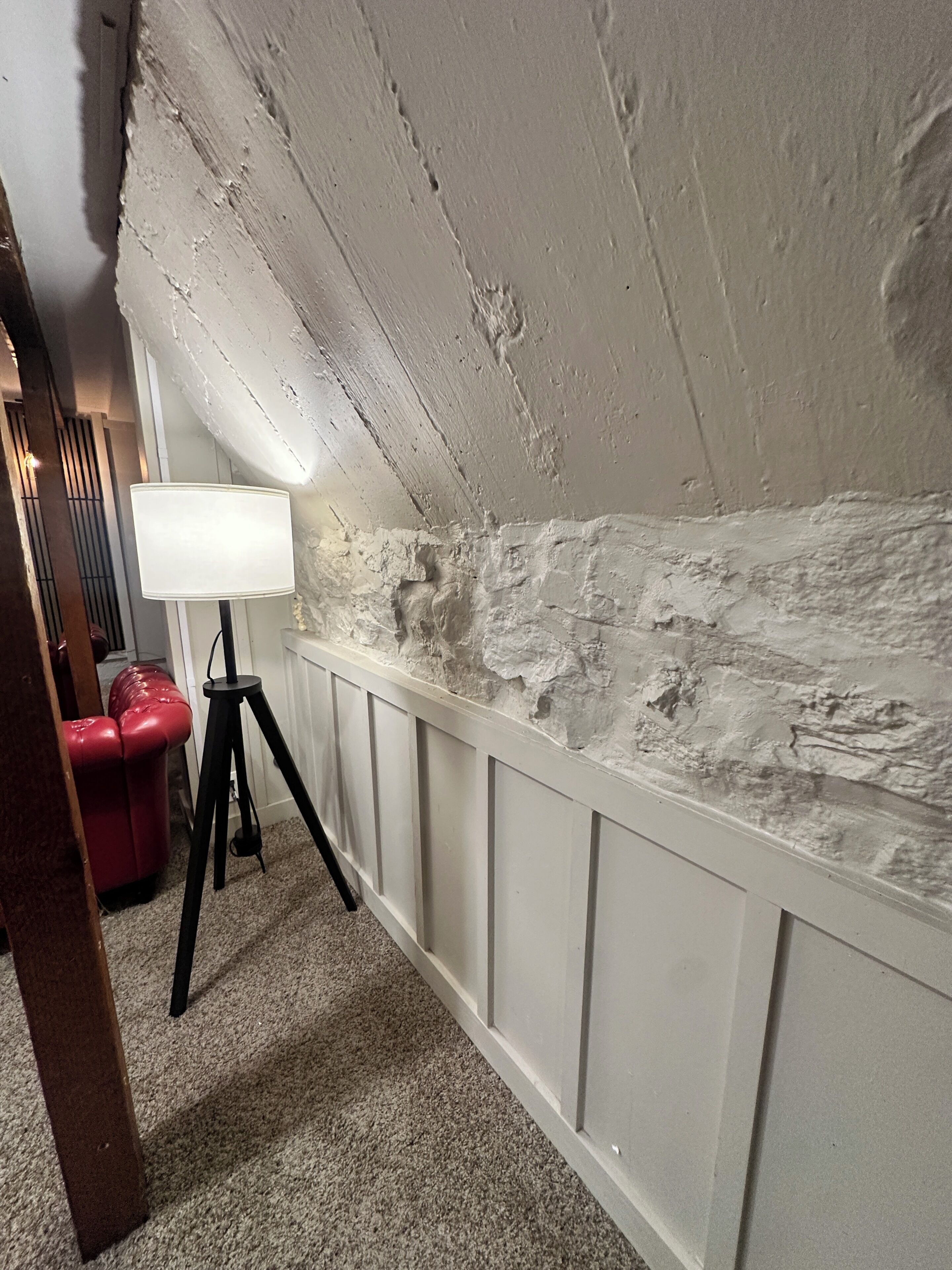 Een zolderkamer met een schuine kap, een statief met een lamp, een rode stoel en een witte muur met een structuur.