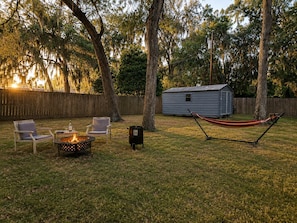 A backyard with a fire pit, two chairs, a hammock, a shed, and a wooden fence.