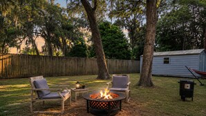 A backyard with a fire pit, two chairs, a table with a lantern, and a grill. - The Vibe House - 3BR Island Retreat near Savannah & Tybee (Savannah)